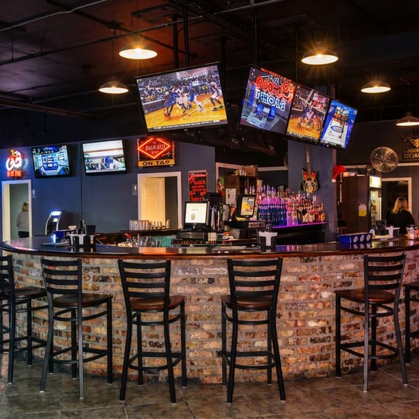Lively bar with a brick counter, patrons, pool players, and several screens displaying sports.