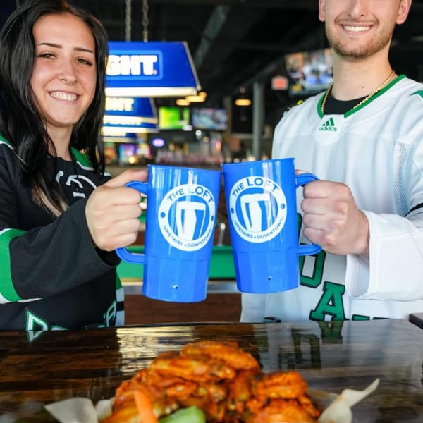 Two smiling people in sports jerseys clinking blue mugs above a basket of chicken wings.