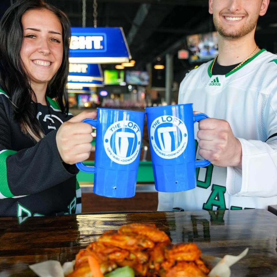Two smiling people in sports jerseys clinking blue mugs above a basket of chicken wings.