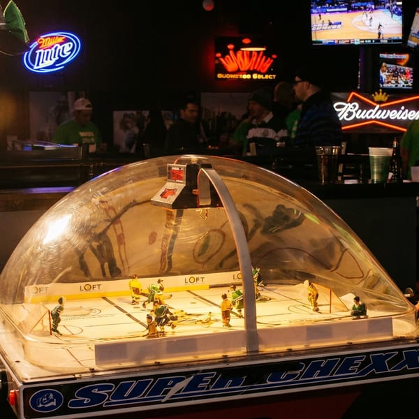 Two men playing Super Chexx bubble hockey in a dimly lit bar with neon signs.