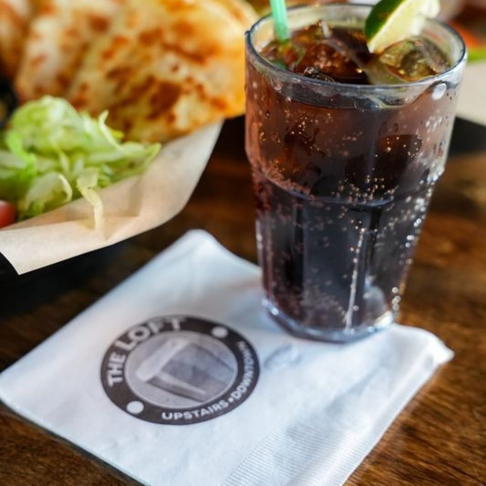 A glass of soda with lime and straw, next to quesadillas and a "The Loft" napkin.