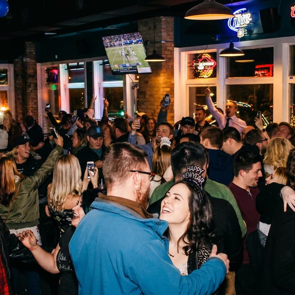 A lively crowd of people celebrating inside a bar with balloons, TVs, and neon signs.