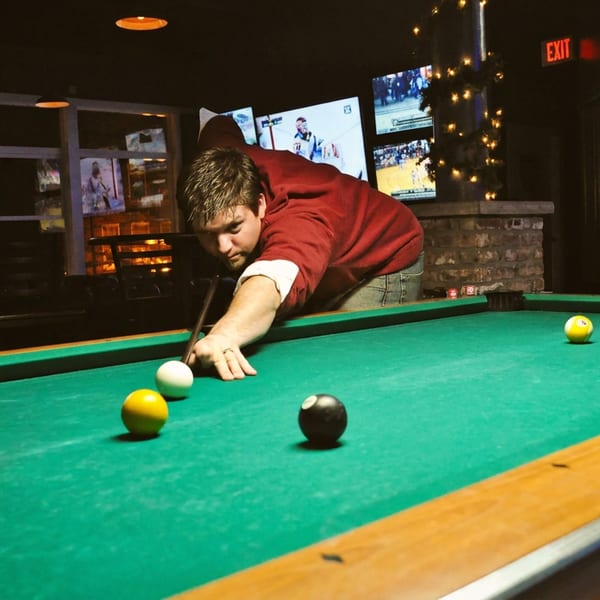 Man playing pool on a green table in a dimly lit bar with TVs in the background.