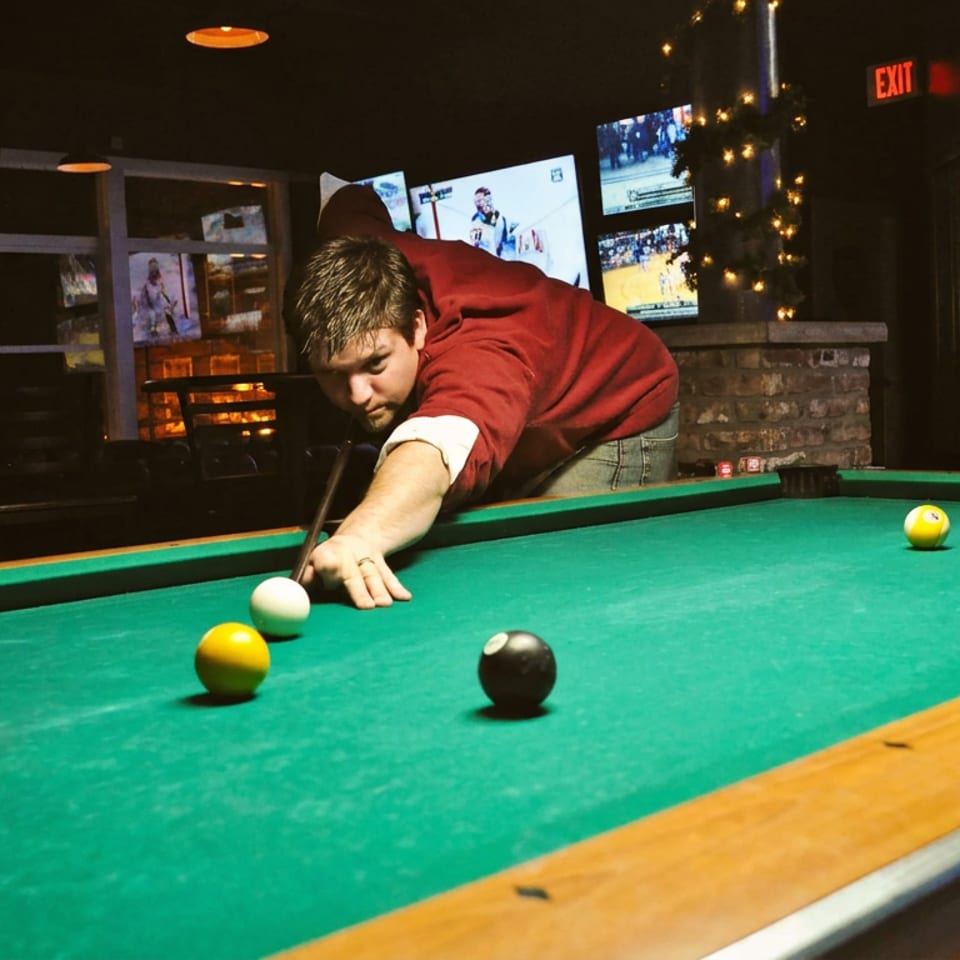 Man playing pool on a green table in a dimly lit bar with TVs in the background.