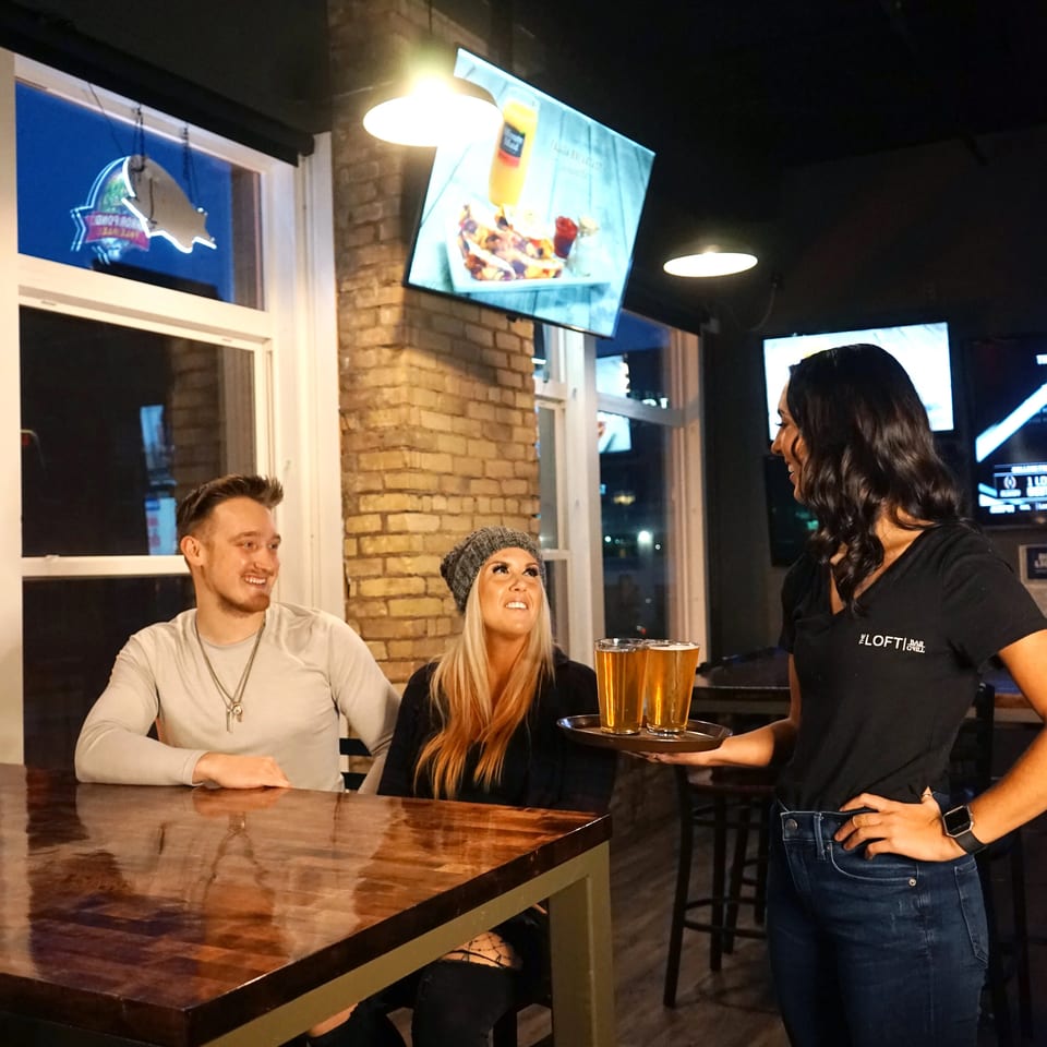 Waitress from The Loft Bar & Grill serves two beers to a couple at a table.