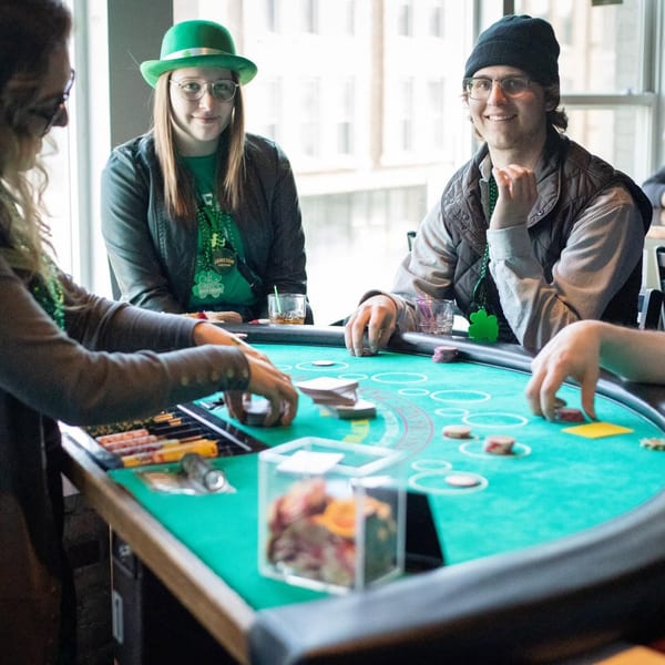 Four people in green attire playing cards at a casino table, celebrating St. Patrick's Day.