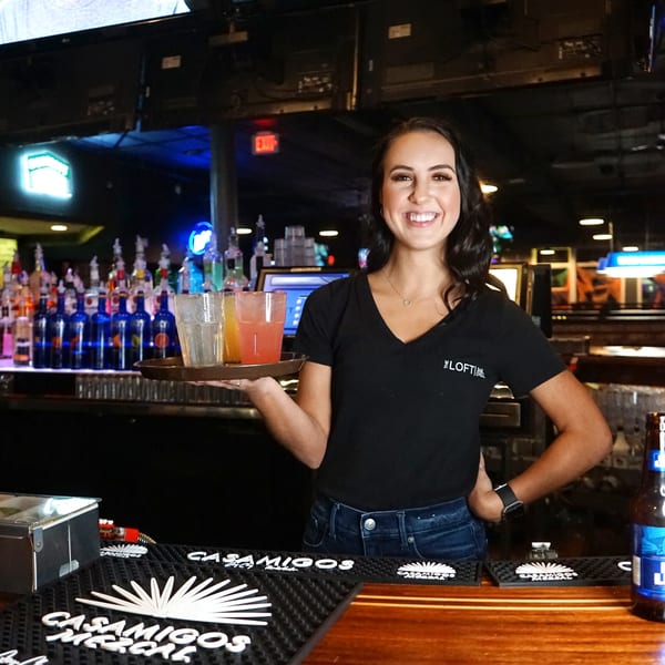 Smiling bartender at The Loft Bar & Grill holds a tray of cocktails behind a well-stocked bar.