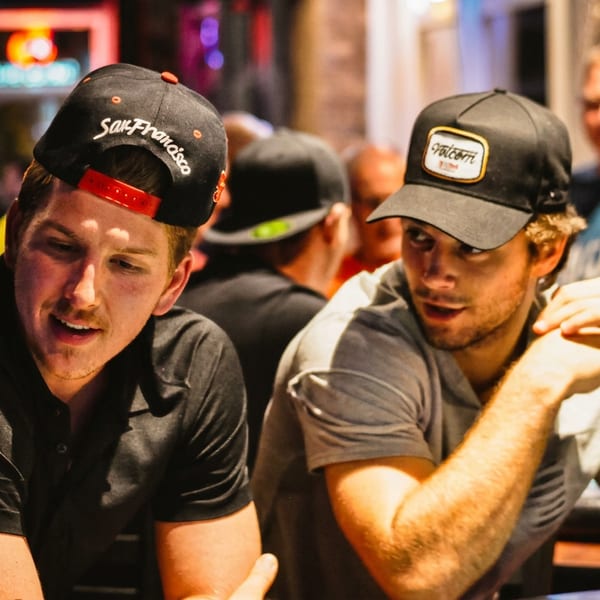 Two men in hats conversing at a lively bar with other patrons in the background.