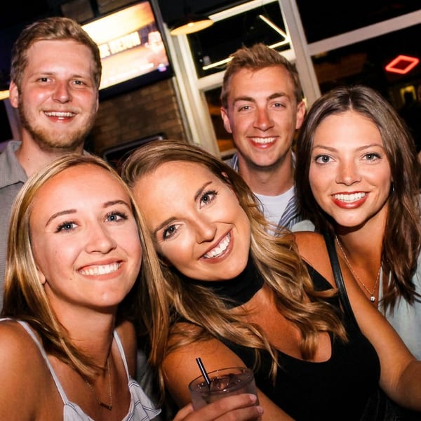 Five smiling friends enjoying drinks together at a vibrant bar with neon signs.