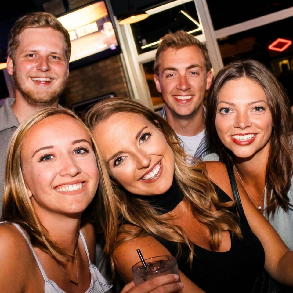 Five smiling friends enjoying drinks together at a vibrant bar with neon signs.