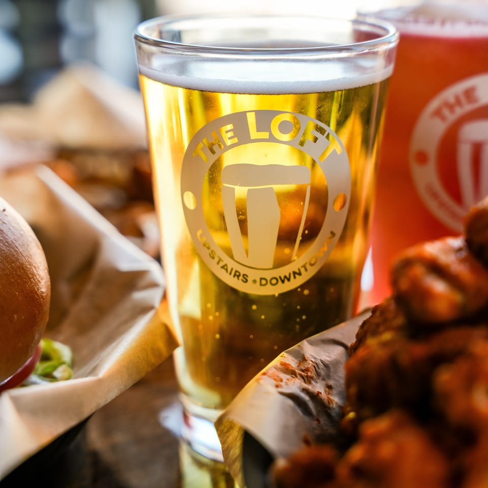 A pint glass of golden beer with "The Loft" logo, next to a burger and chicken wings.