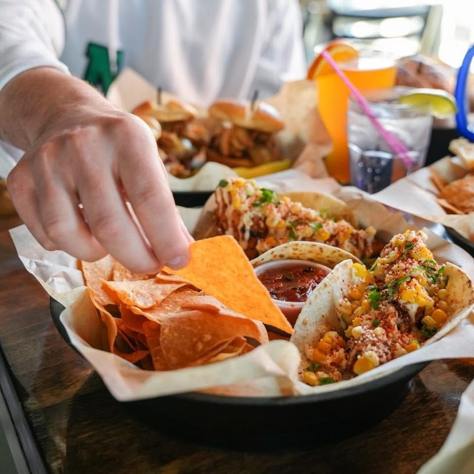 A person's hand reaches for a chip from a basket of tacos, chips, and salsa.