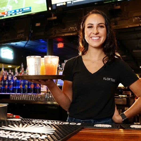 Smiling bartender at The Loft holding a tray with two drinks, with sports on TVs in the background.