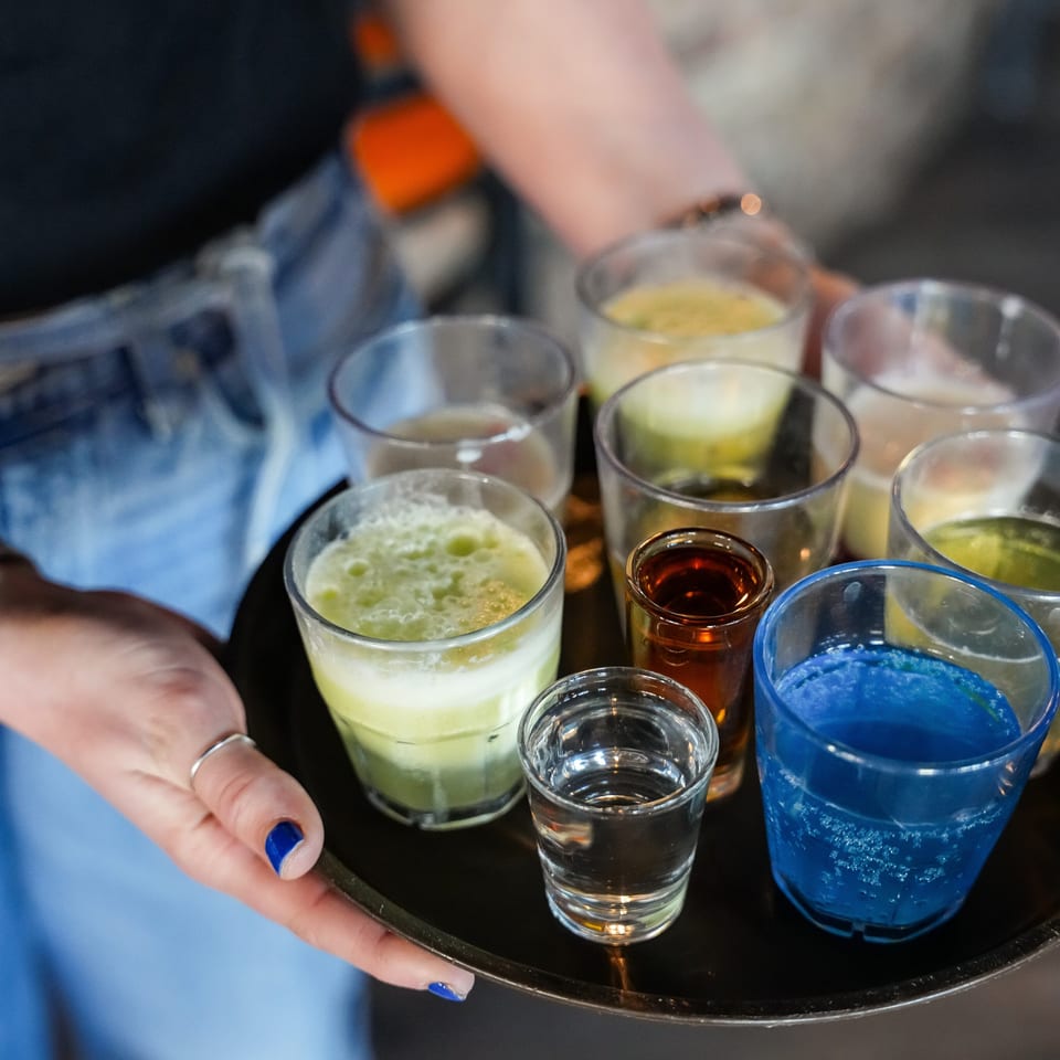 A person holds a black tray with nine small glasses of colorful drinks, including green, blue, and amber.