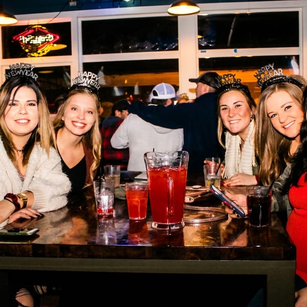 Five smiling women wearing "Happy New Year" headbands at a bar table with drinks.