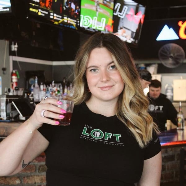 Smiling woman in a black "The Loft" shirt holding a red drink in a lively bar setting.