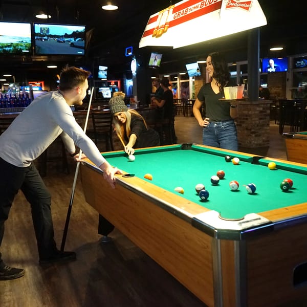 Man and woman playing pool in a bar, with a server holding drinks.