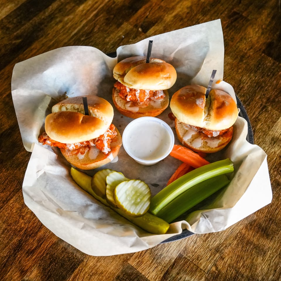 Three buffalo chicken sliders with ranch, celery, carrots, and pickles in a basket.