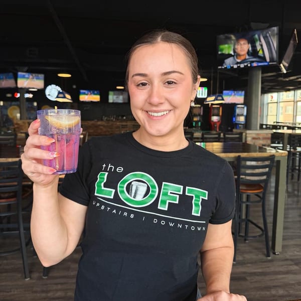 Smiling woman in a black "the LOFT" t-shirt holding a pink drink with lemon in a bar.